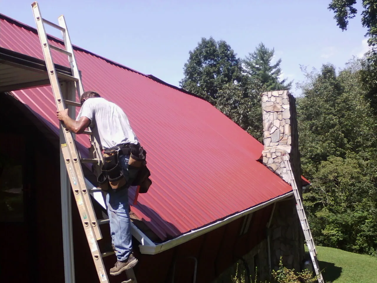 Roofer on a ladder beside a red metal roof and stone chimney — Soffit & Fascia Repair by Roofing Brothers in Duchesne